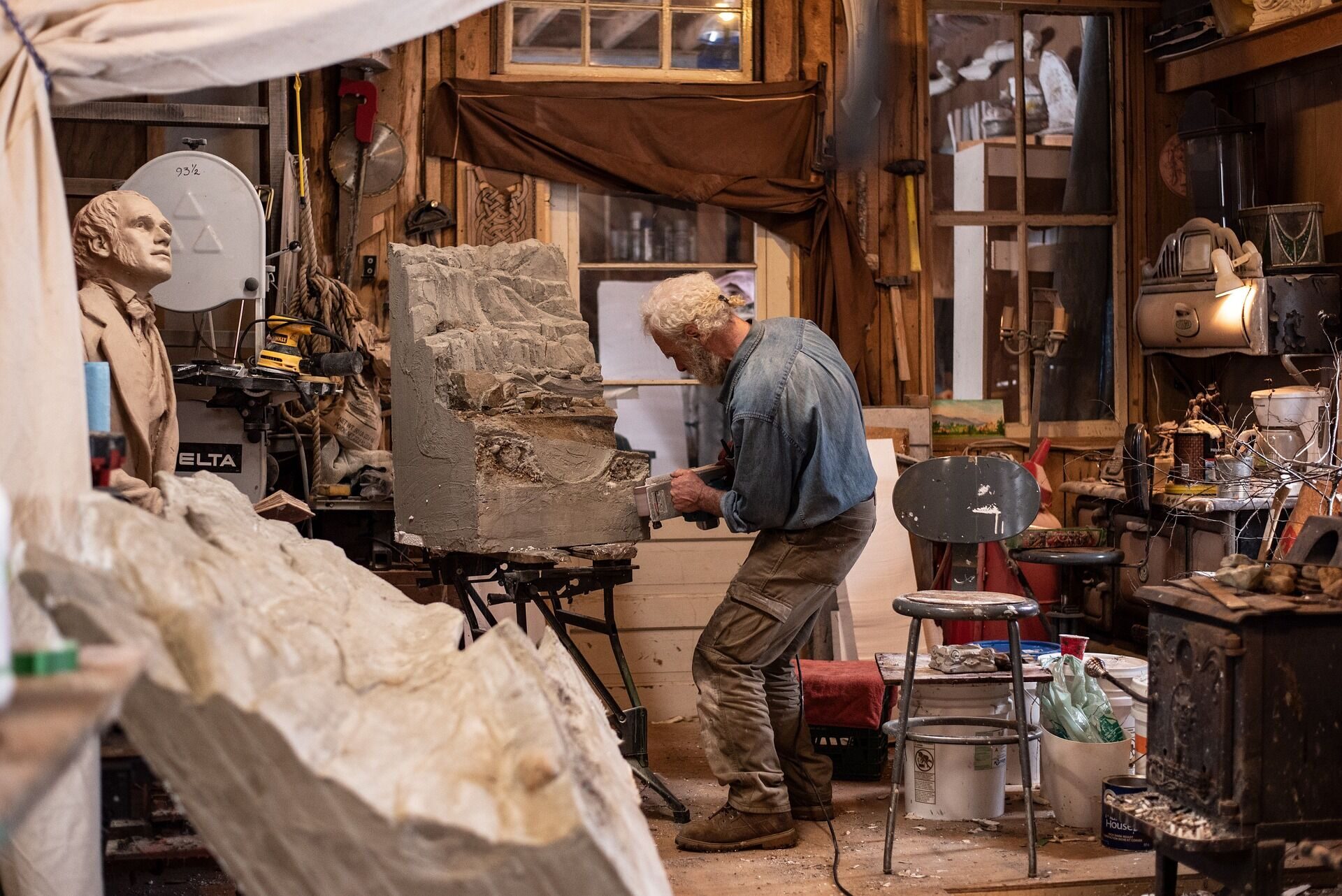 A sculptor carves detail into a large clay relief in a cluttered studio workshop.